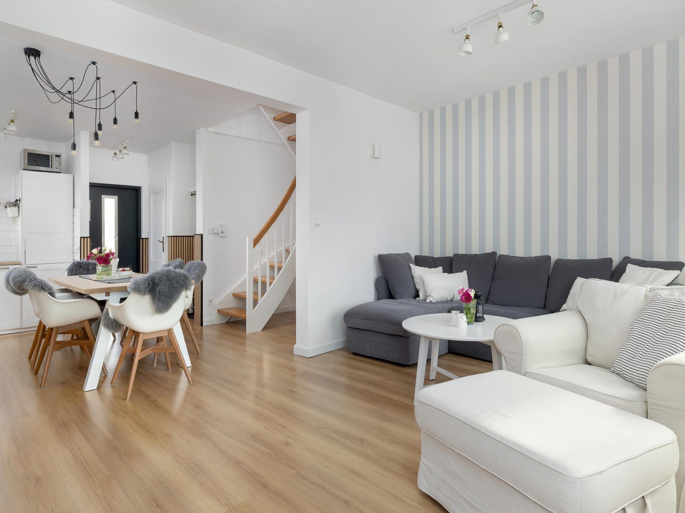 A modern living room with a grey sofa, white armchairs, a wooden dining table with chairs, a staircase, and a striped wall.