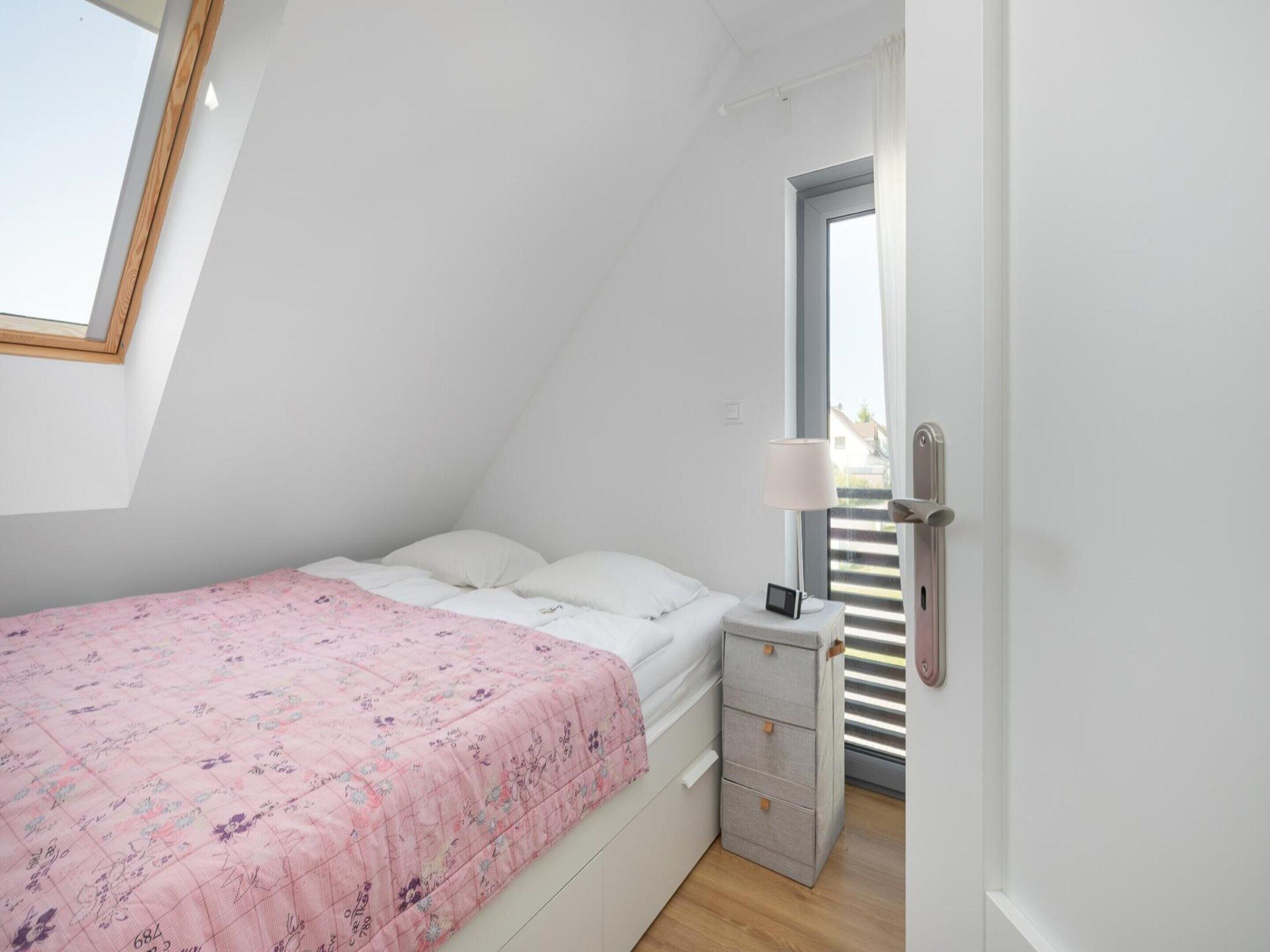 Una habitación pequeña con cama individual, techo inclinado, ventana con persianas, mesita de noche con lámpara y un mueble para guardar objetos.