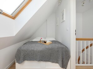 A bedroom with a sloped ceiling, a bed with a gray blanket, a bedside table with a book and a vase, and a white railing. - Holiday Home, 3 Bedrooms, Private Garden (Rewal)