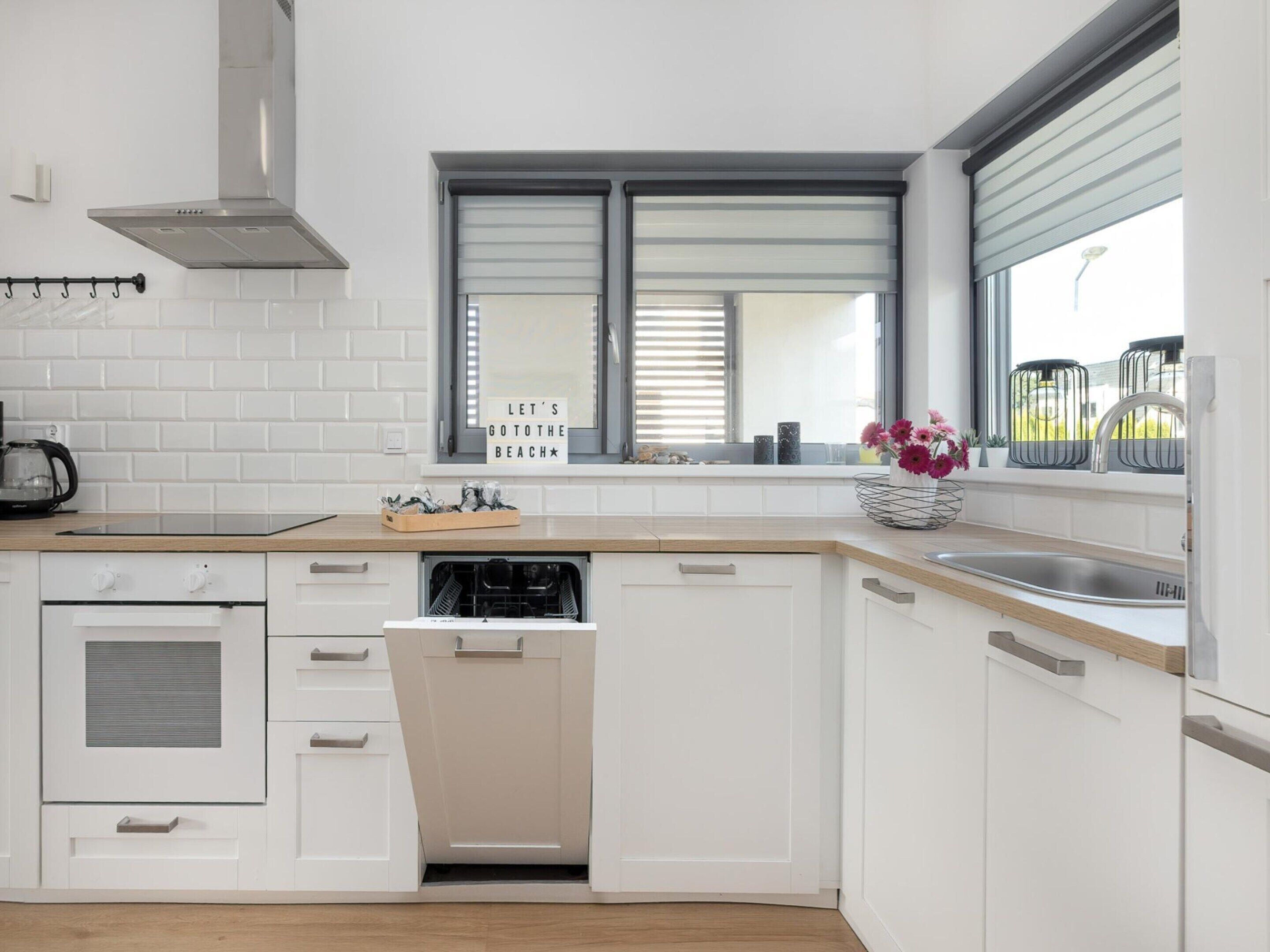 Une cuisine moderne avec des meubles blancs, un lave-vaisselle, une cuisinière et une fenêtre équipée de stores.