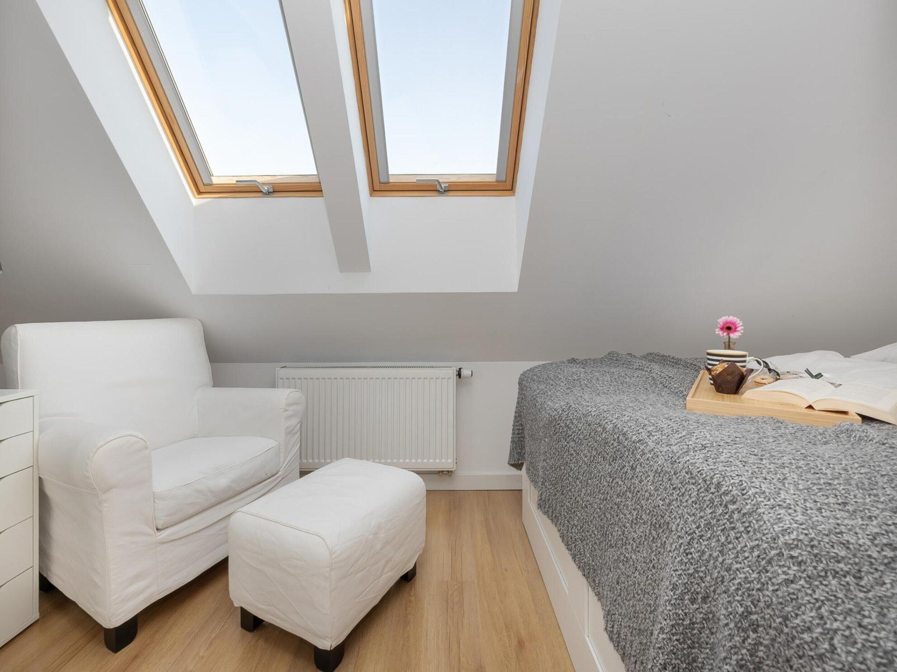 A bedroom with a skylight, a bed with a grey blanket, a white chair, and a small bedside table with a pink flower.