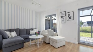 A modern living room with a grey sofa, a white round table, and a white armchair. There are striped wallpaper and a large window with curtains.