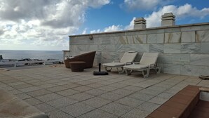 A rooftop terrace with a view of the sea, featuring a stone wall, lounge chairs, and a wicker chair. - Luxury Apartment with Private Pool on Huge Terrace- Ideal for Families and Expats (Las Palmas de Gran Canaria)