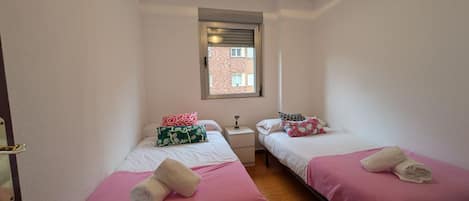 A small, clean room with two beds, a window, a nightstand, and a door.