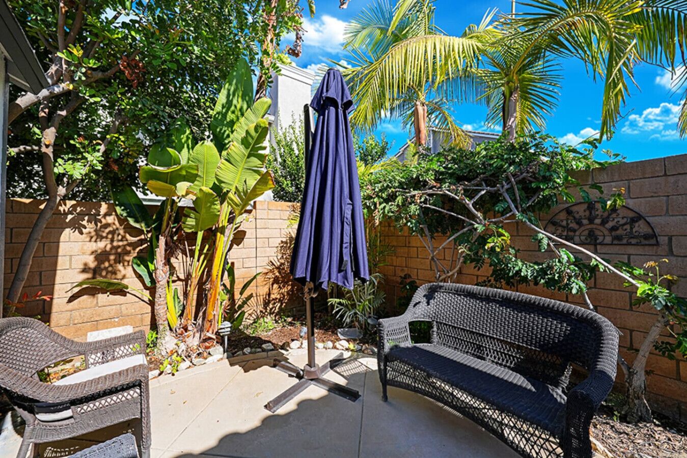 A patio area with a wicker bench, a blue umbrella, and lush greenery including banana plants.