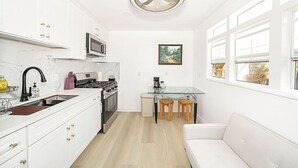 Una cocina moderna con armarios blancos, microondas integrado, estufa a gas y ventilador de techo. Hay un comedor con mesa de vidrio y dos taburetes de madera.
