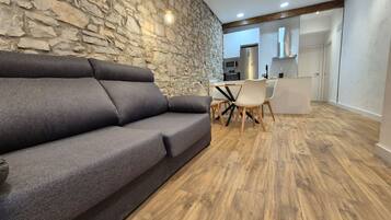 A modern living room with a stone wall, a grey sofa, a wooden dining table with chairs, and a kitchen in the background.