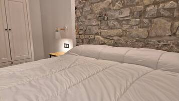 Un dormitorio con pared de piedra, una cama con ropa de cama blanca, una mesita de noche de madera y un armario blanco.