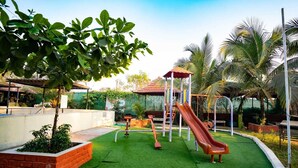 A playground with a slide and climbing structures, surrounded by green grass and palm trees. - Mathura Resort (Alibag)