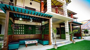 A covered outdoor seating area with a bench and table, surrounded by potted plants and a green lawn. - Mathura Resort (Alibag)