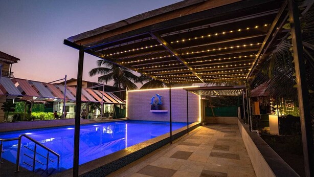 A swimming pool with a covered area and string lights, surrounded by a brick wall and palm trees. - Mathura Resort (Alibag)