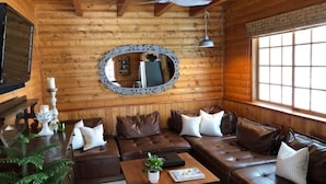 Interior de una cabaña de troncos con techo de madera, un ventilador de techo, un espejo grande, sofás de cuero, una mesa de centro y una ventana con persianas.