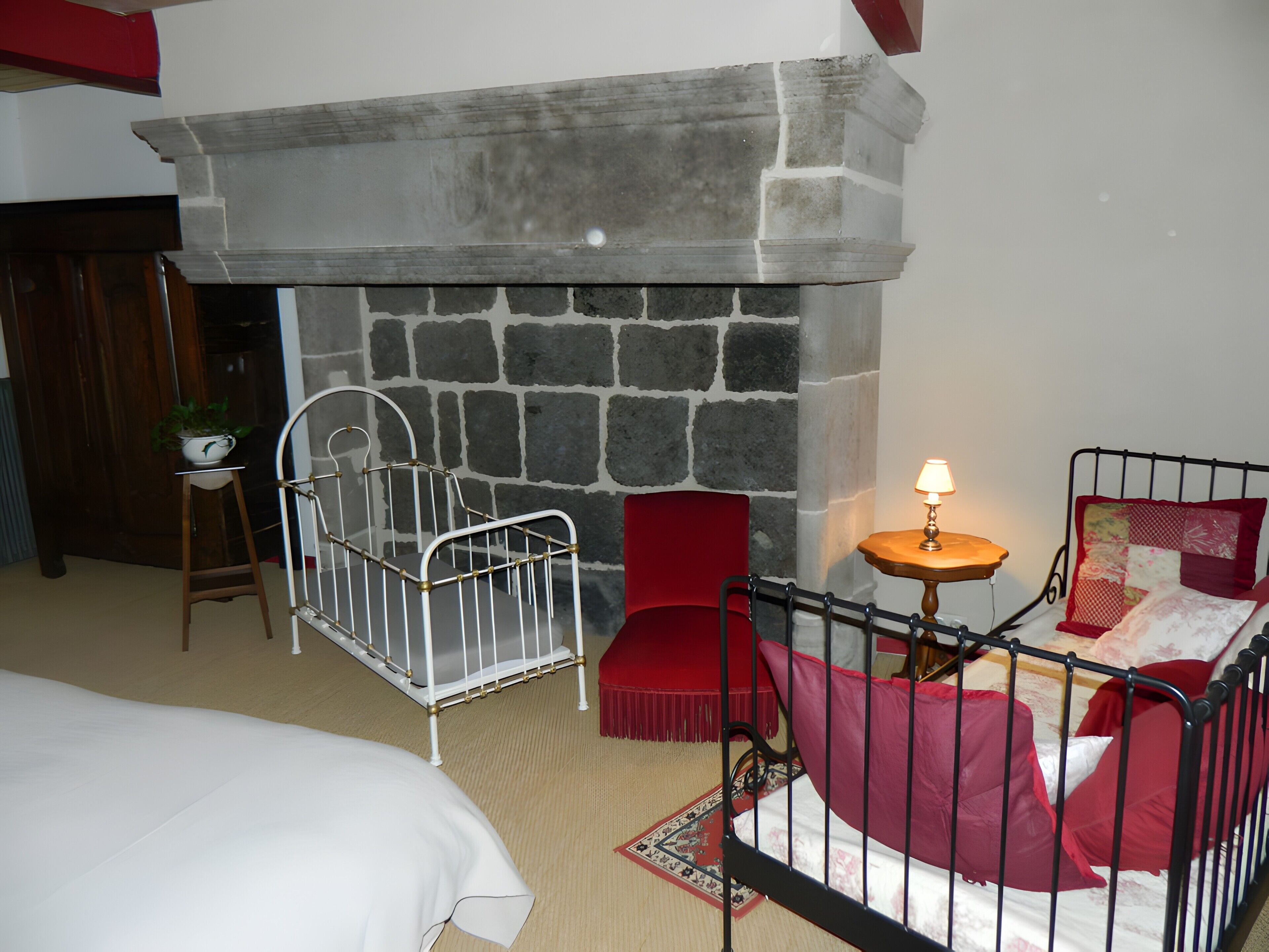Ein Zimmer mit Kamin aus Stein, einem weißen Babybett, einem roten Sessel, einem kleinen Tisch mit Lampe und einem Bett mit weißer Bettdecke.
