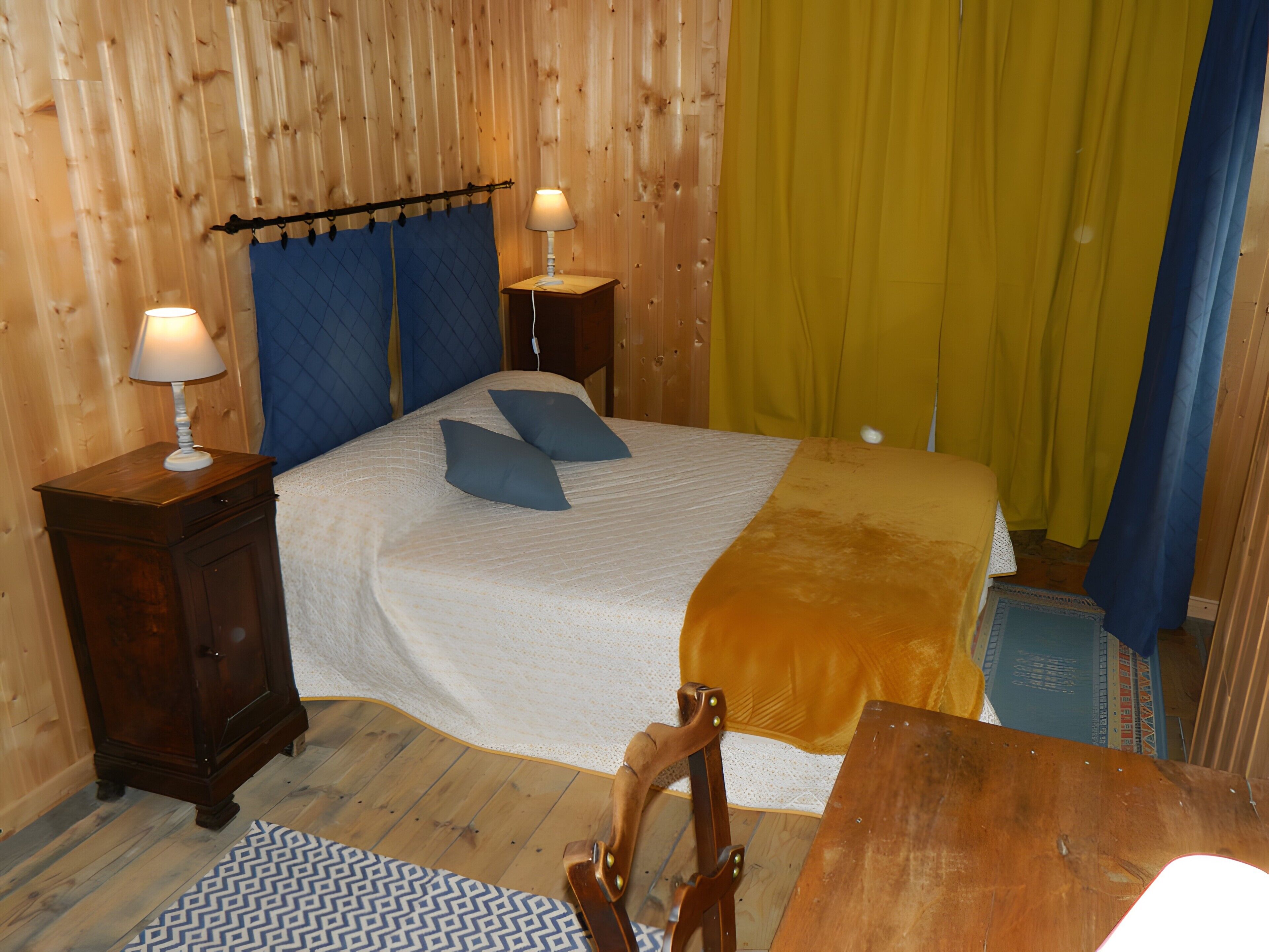 A wooden-paneled room with a bed, two bedside tables with lamps, a wooden chair, and a small table.
