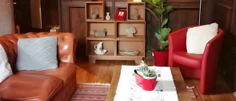 Una sala de estar acogedora con un sofá de cuero, un sillón rojo, una mesa de centro con una planta en maceta, estanterías de madera con objetos decorativos y una lámpara de pie.