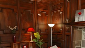 A room with wooden paneling, a red armchair, a sofa, a lamp, a potted plant, and a small table with a placemat.