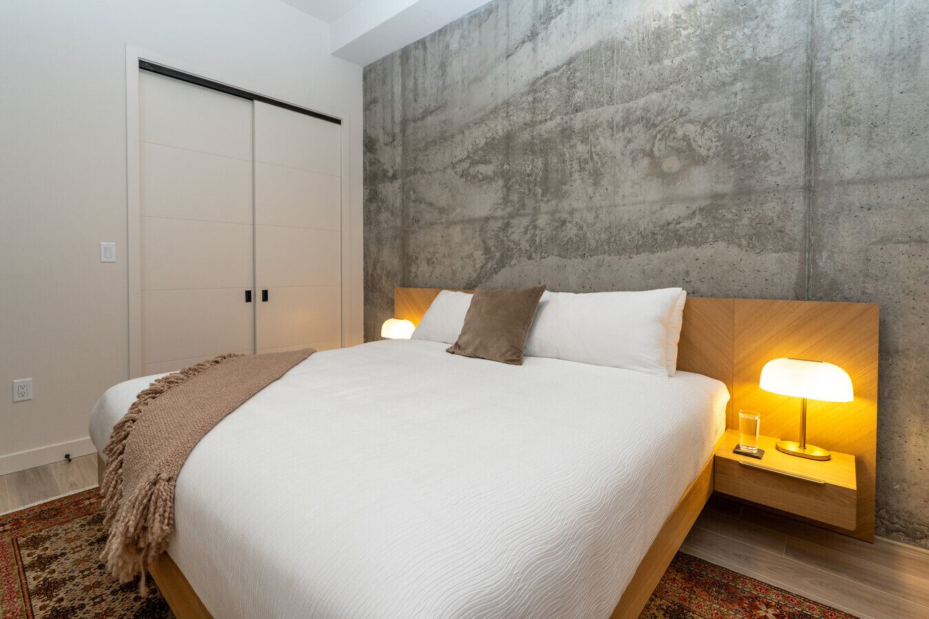 Une chambre à coucher moderne avec un grand lit, un mur d’accent en béton et une table de chevet en bois avec une lampe.