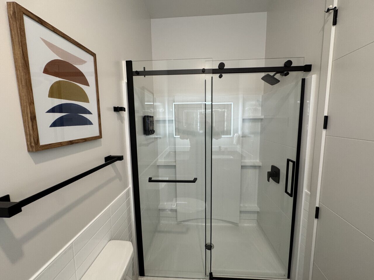 Une cabine de douche fermée par des parois de verre, avec un mur recouvert de carreaux blancs, une œuvre d’art abstraite encadrée au-dessus de la porte et un porte-serviettes noir.