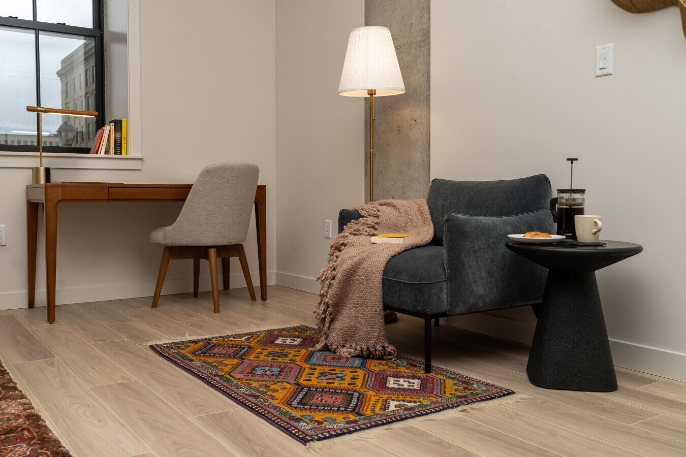 Een moderne woonkamer met een grijze bank, een klein rond tafeltje met een kop en theepot, een houten bureau met een stoel, en een kleurrijk vloerkleed.