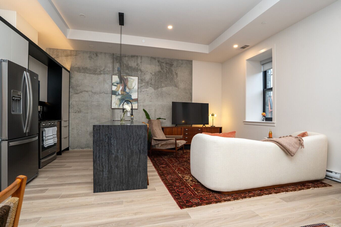A modern kitchen with a built-in refrigerator, oven, and a central island with a sink. There is a flat-screen TV on a wooden cabinet, a white sofa, and a red patterned rug.