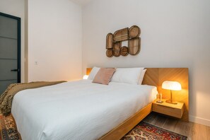 A neatly made bed with a white comforter and pillows, a wooden headboard, a wall-mounted decorative piece, a bedside lamp, and a patterned rug. - *NEW Mid-Century Modern 2B/1B (Halifax)