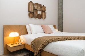 A modern bedroom with a wooden headboard, a bedside lamp, a bed with white bedding and a brown throw blanket, and a wall-mounted abstract art piece.