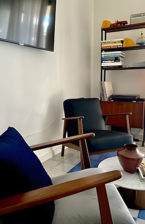 A modern living room with a flat-screen TV, a blue armchair, a wooden side table, a bookshelf with books and decorative items, and a small table with a vase.