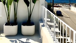 Un balcó amb plantes en testos, una barana blanca i vista al mar i a la carretera.