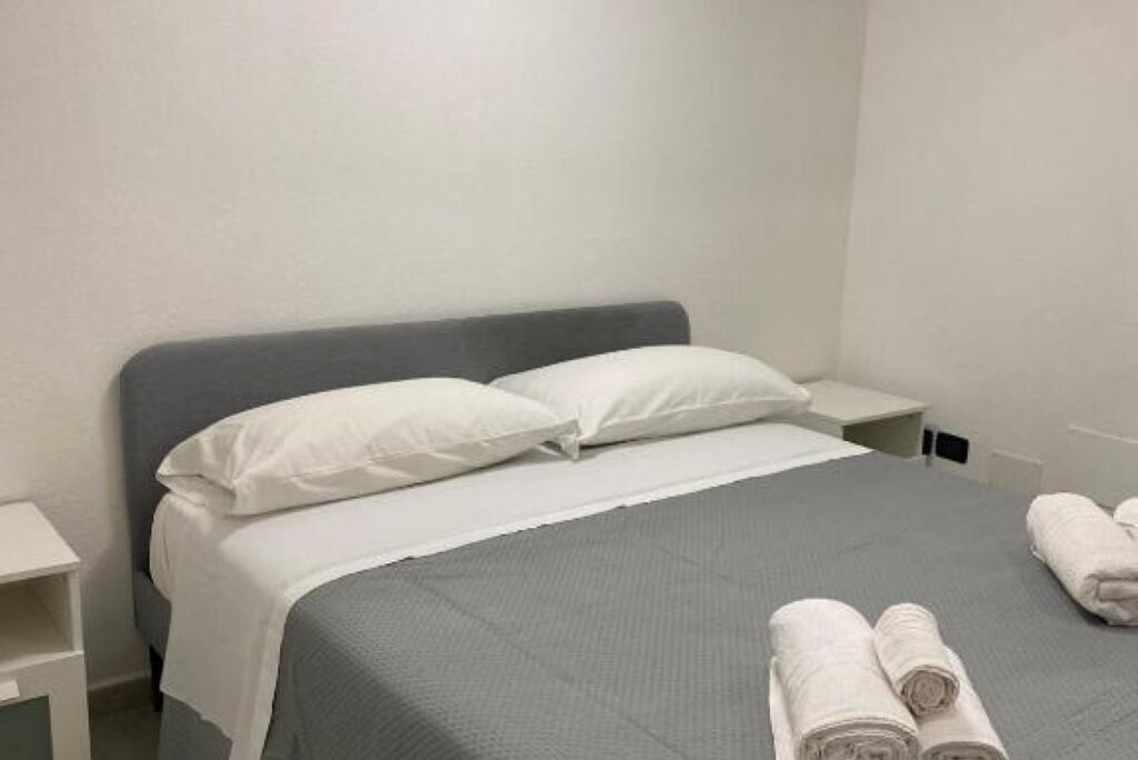 A neatly made bed with white bedding and pillows, a grey headboard, and two rolled towels on the side.
