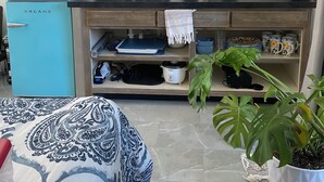 A compact kitchen with a blue refrigerator, a microwave, a kettle, and a sink. There are shelves with dishes and a potted plant.