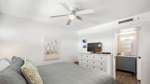 A bedroom with a bed, a ceiling fan, a TV, a dresser, and a bathroom visible through an open door.