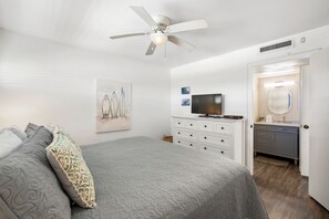 Ein Schlafzimmer mit Bett, Deckenventilator, Fernseher, Kommode und einem durchgegangene Tür sichtbaren Badezimmer.
