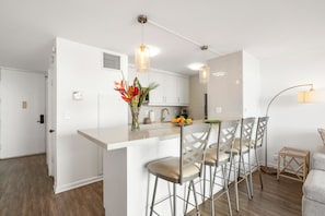 Une cuisine moderne avec des meubles blancs, un îlot central doté de tabourets, des luminaires suspendus et un vase rempli de fleurs.