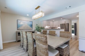 A dining area with a long table, wicker chairs, a chandelier, and a view of the kitchen.