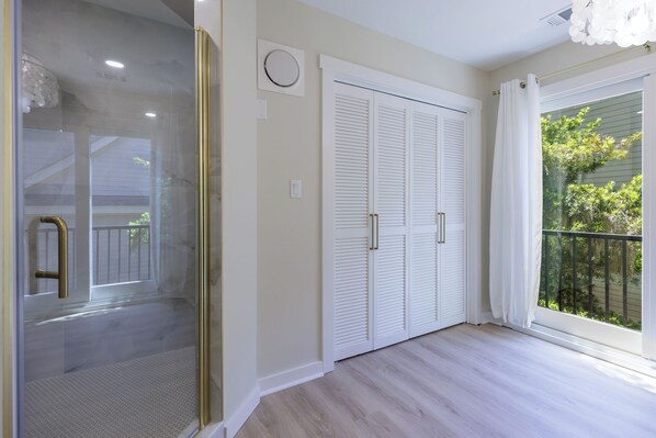 A room with a glass door leading to a balcony, a set of white louvered closets, and a large window with a view of greenery.