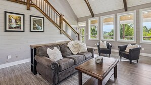 A spacious living room with a high ceiling, a wooden staircase, and large windows offering a view of the outdoors. - Amazing Views Hot Tub Arcade Firepit (Gatlinburg)