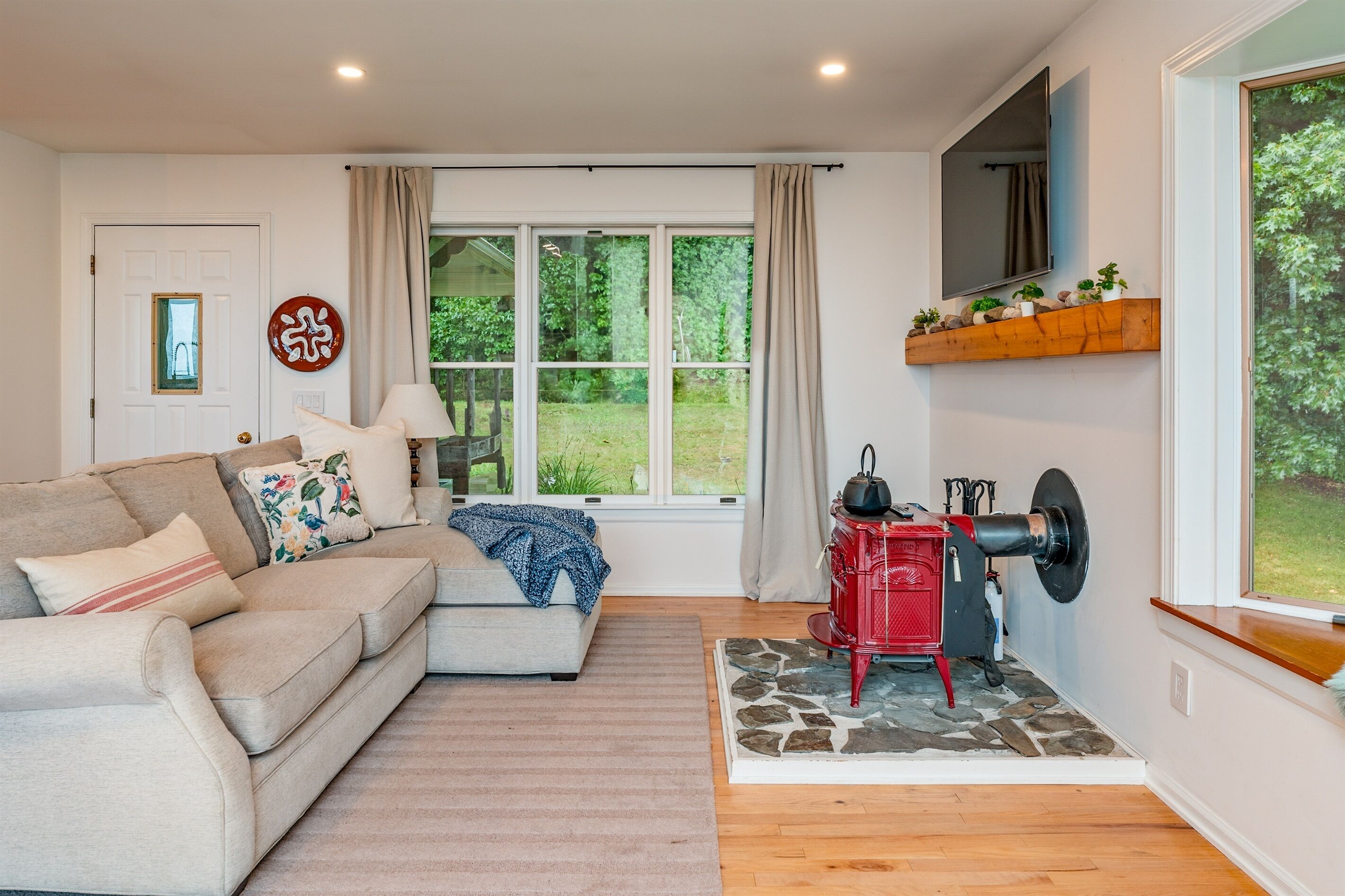 A cozy living room with a sofa, a red stove, a large window with curtains, and a stone-patterned rug.