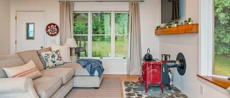 A cozy living room with a sofa, a red stove, a large window with curtains, and a stone-patterned rug.