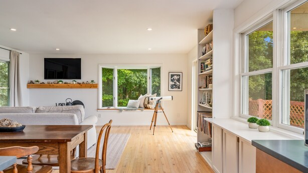 Un salon moderne avec un canapé, une table à manger en bois et des chaises. Une grande fenêtre offre une vue sur la verdure à l’extérieur.