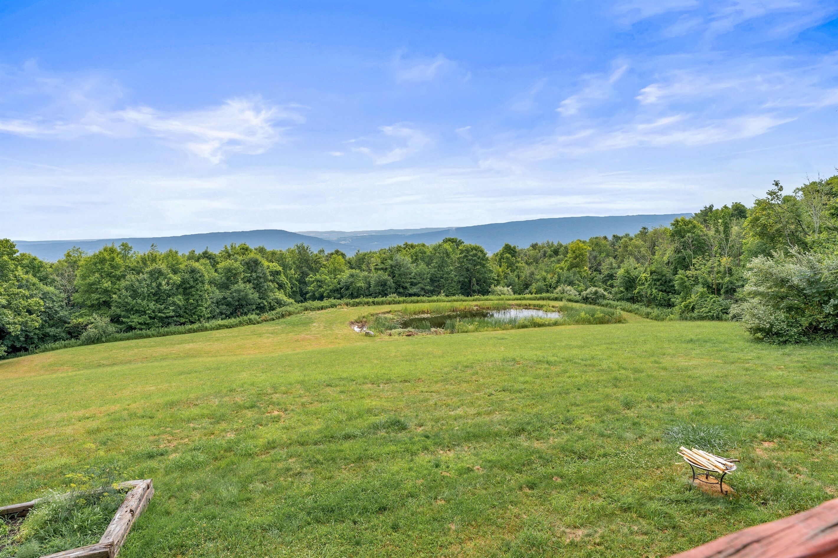 A grassy field with a pond and a bench, surrounded by trees and hills in the distance.
