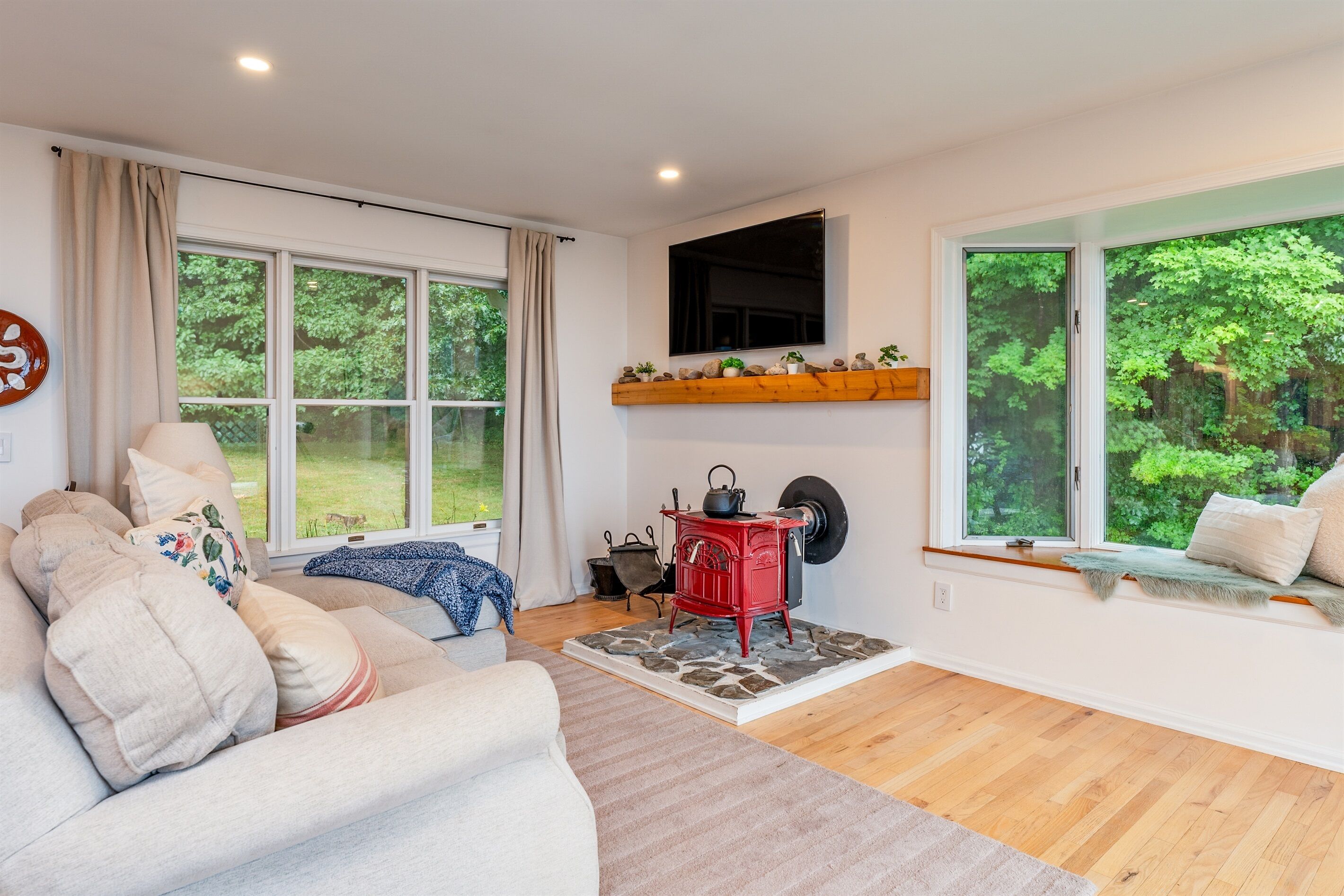 Una sala de estar acogedora con un sofá, una estufa roja y grandes ventanas con vistas al verdor.