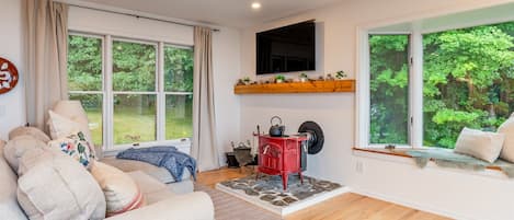 Una sala de estar acogedora con un sofá, una estufa roja y grandes ventanas con vistas al verdor.