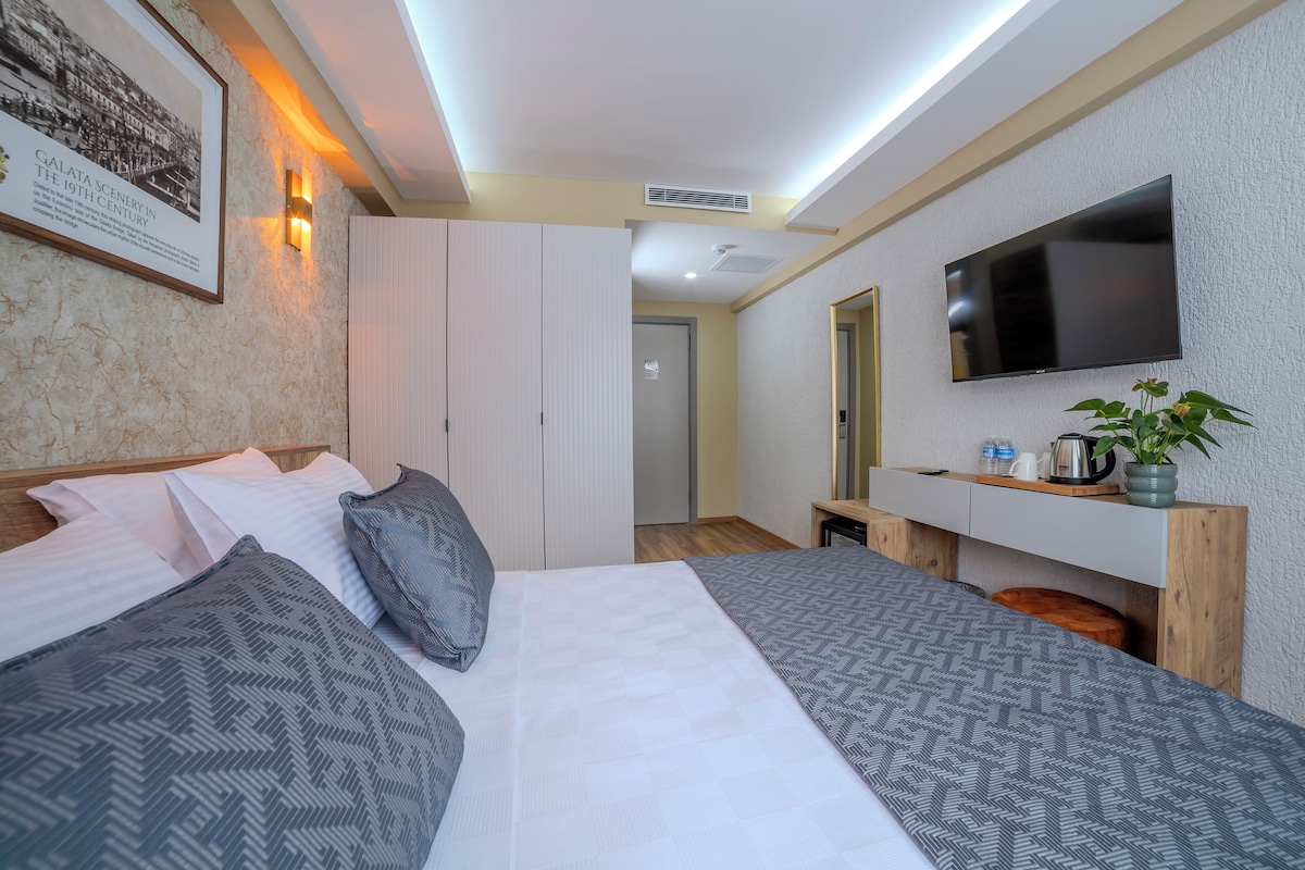 A hotel room with a large bed, a TV mounted on the wall, a small desk with a plant, and a framed picture on the wall.