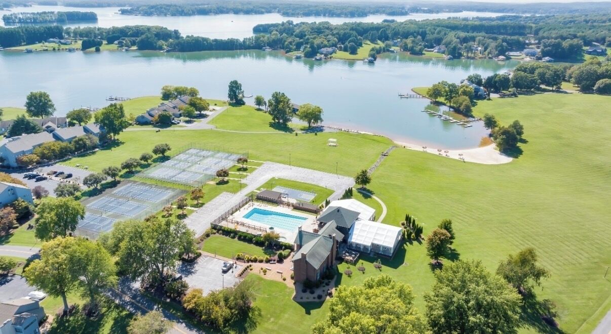 Luftaufnahme eines Sees mit einem Anwesen, das einen Swimmingpool, Tennisplätze und ein Gewächshaus umfasst.