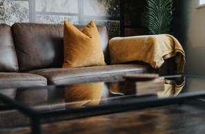 A brown leather sofa with two cushions, a glass coffee table, and a large green plant in the background. - Victoria Place (Hyde)