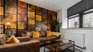 Un salon avec un canapé en cuir marron, des coussins jaunes, une table basse en verre et un mur orné d’un motif en mosaïque.