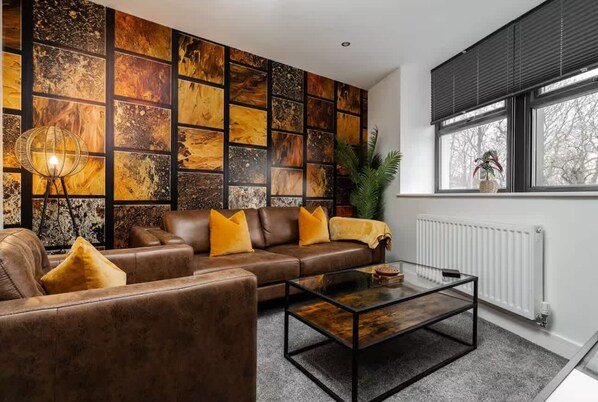 A living room with a brown leather sofa, yellow cushions, a glass coffee table, and a wall with a mosaic pattern. - Victoria Place (Hyde)