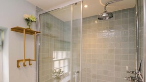 A glass-enclosed shower with a handheld showerhead, a glass door, and a shelf with a vase of flowers. - Beech (Newbury)