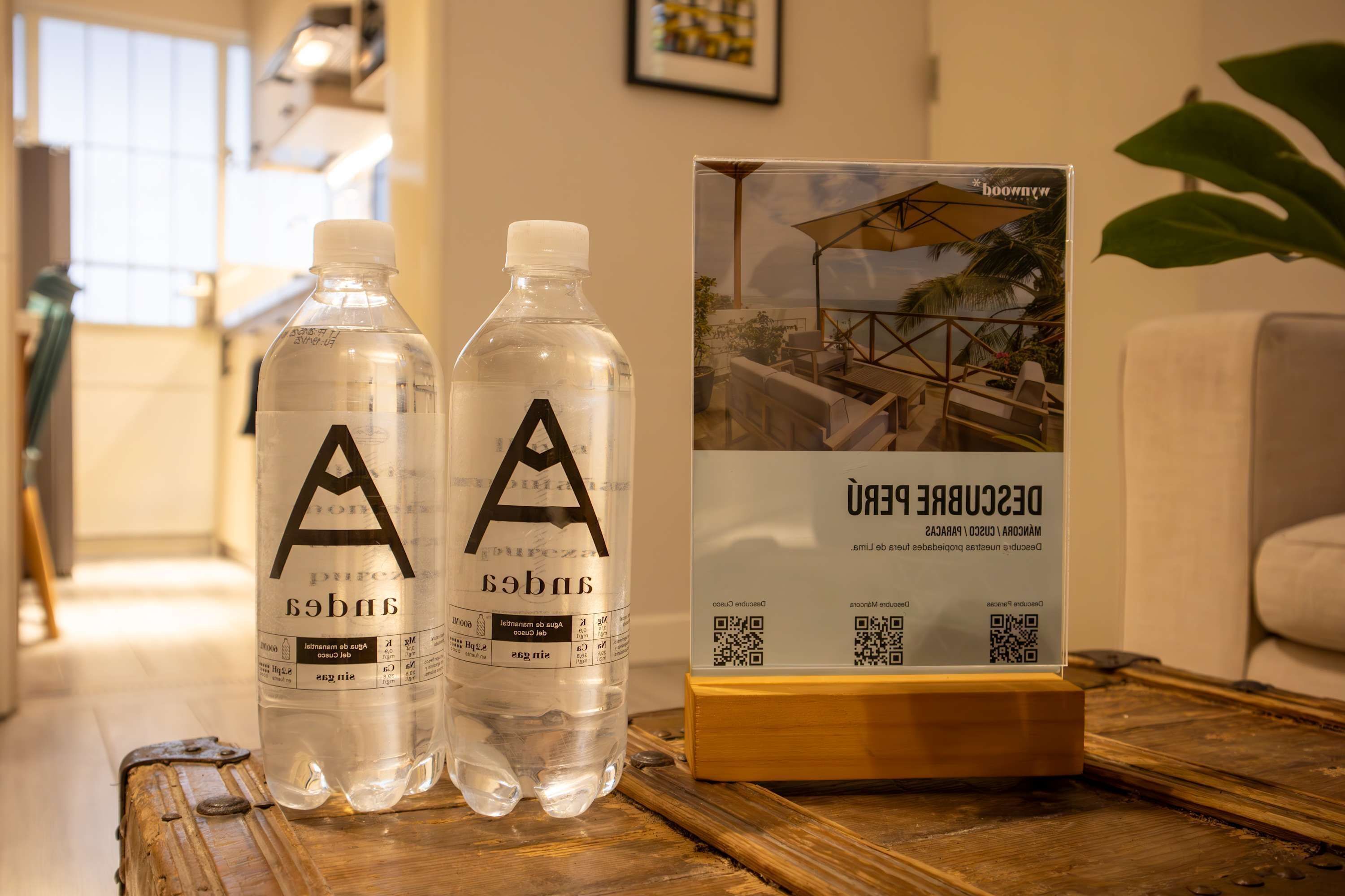 Two water bottles with a logo on a wooden table, a framed picture of a building, and a plant in the background.