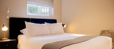 A neatly made bed with white linens and a grey throw blanket, a bedside lamp, and a window with blinds.